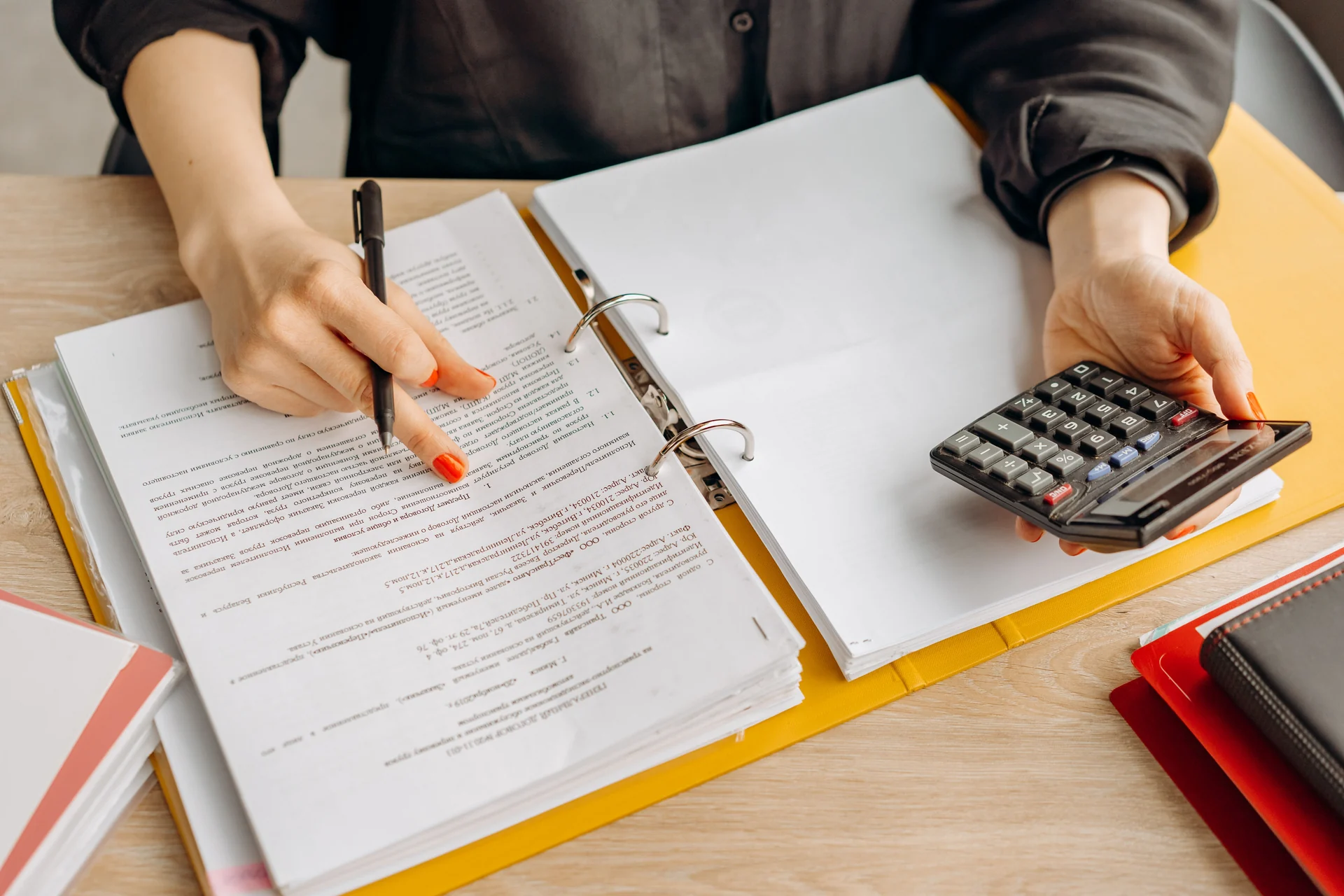 Hands reviewing paperwork in a ring binder while holding a calculator and pen at a desk.